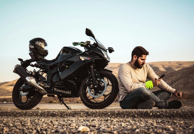 A man sits beside his Kawasaki bike, using a smartphone in a serene outdoor setting.